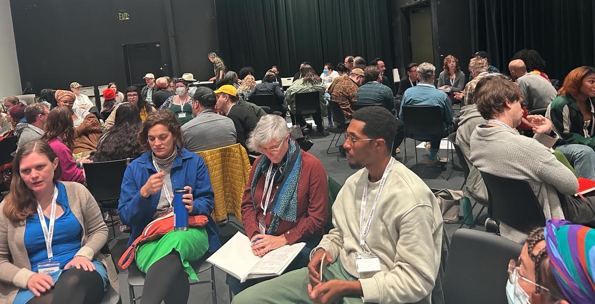 A diverse group of people sit in a large, dark room, attending a workshop or discussion.