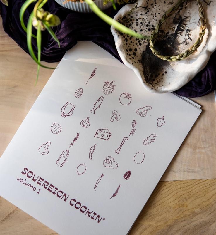 A white cookbook titled "SOVEREIGN COOKIN' volume 1" featuring food illustrations, placed on a wooden table with a glass jar and silverware.