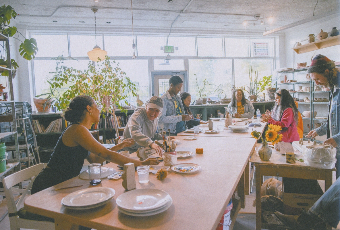 A diverse group of people painting ceramic items at a long table in a bright workshop.