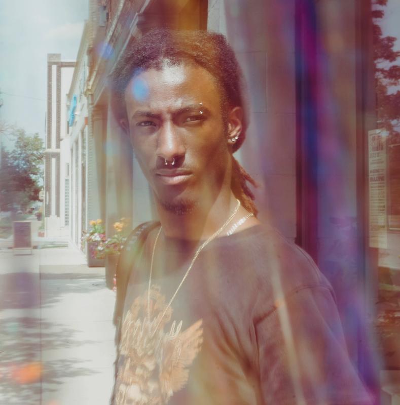 A person with dreadlocks, nose ring, and eyebrow piercing looks forward, seen through colorful lens flares and a soft haze.
