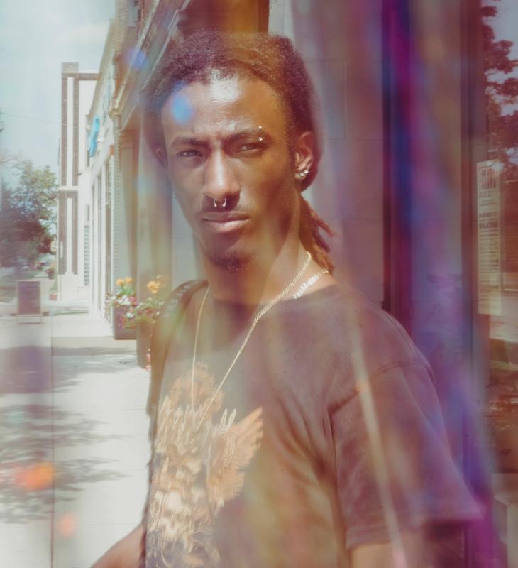 A person with dark skin, dreadlocks, and facial piercings looks directly forward, bathed in colorful lens flares with an urban background.