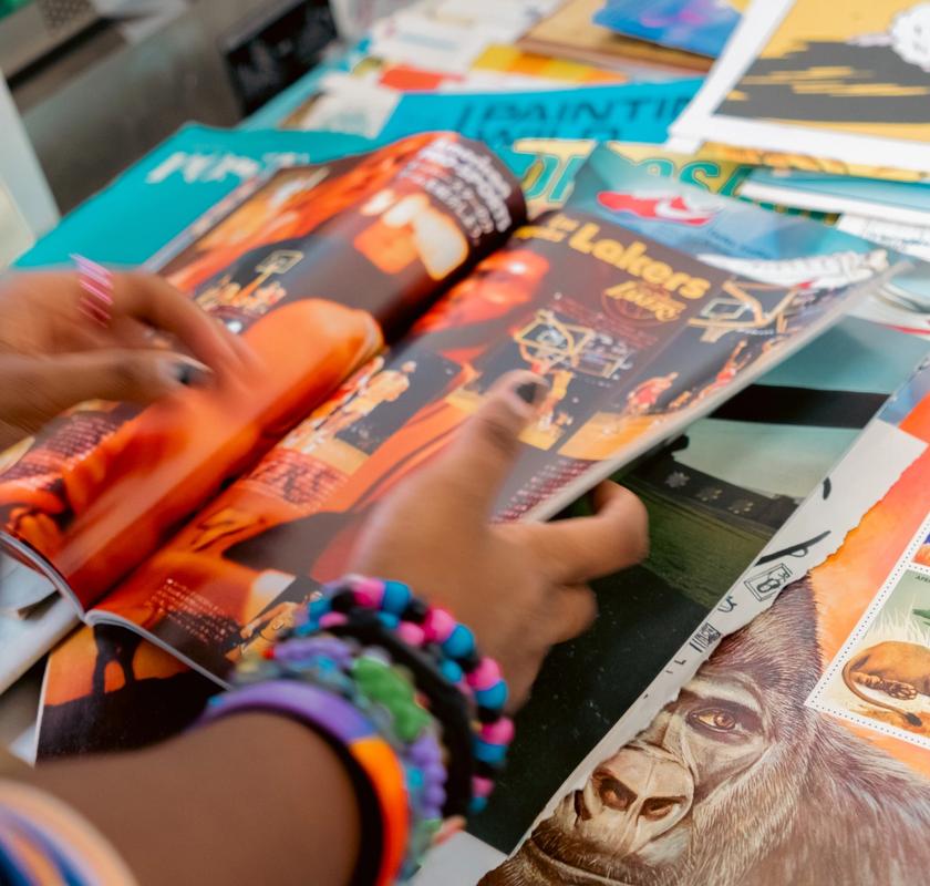 Hands wearing colorful bracelets flip through a magazine, with "Lakers" visible on the page, surrounded by other publications.