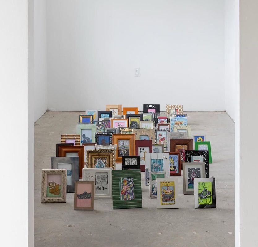 A large collection of diverse framed pictures and artworks are arranged in a rising formation on a concrete floor in a minimalist white room.