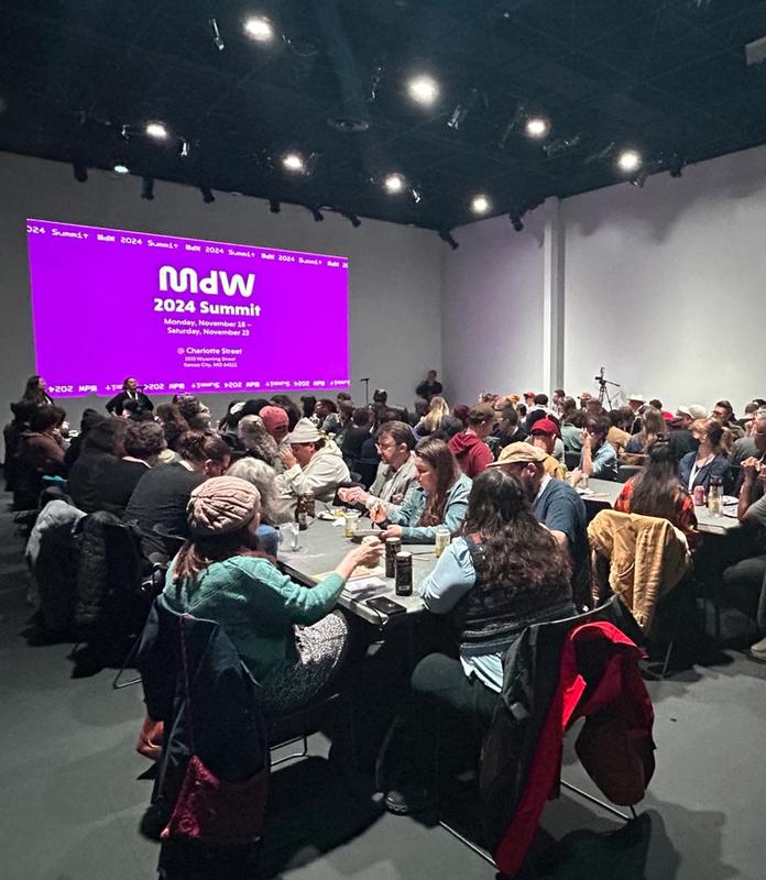 A large group of people seated at tables in a room, facing a screen displaying "MDW 2024 Summit" on a purple background.