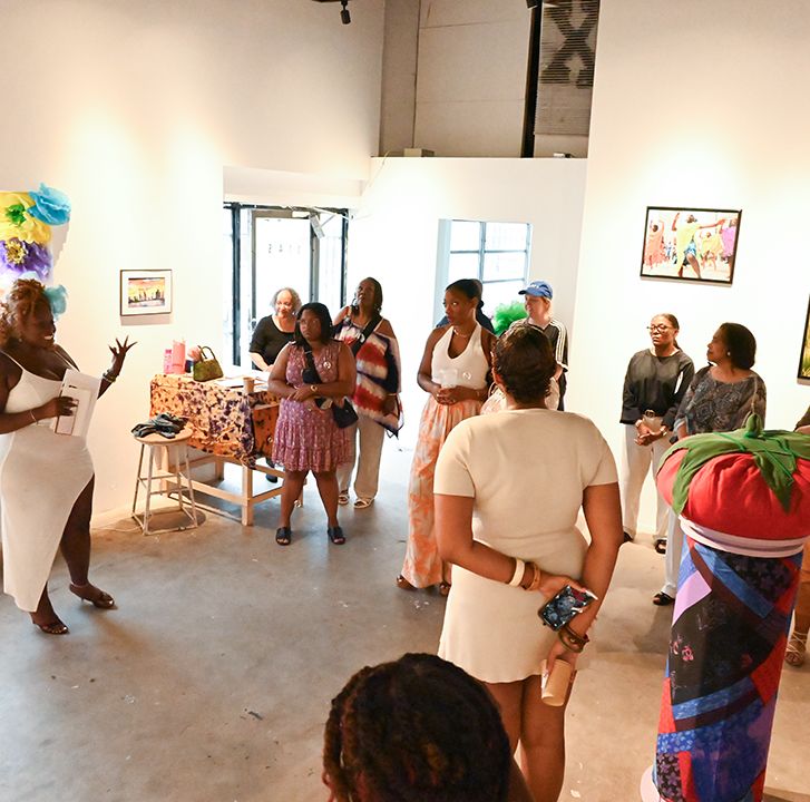A woman in a white dress speaks to a group of people in an art gallery, surrounded by framed artworks and a colorful sculpture.