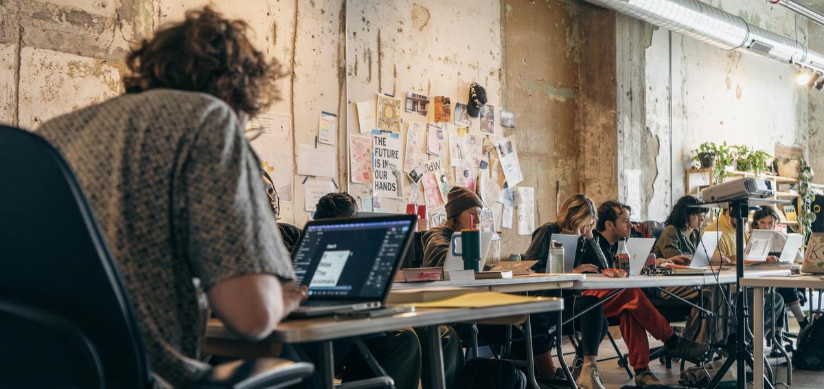 People work on laptops at tables in an industrial-style room with notes on the wall.