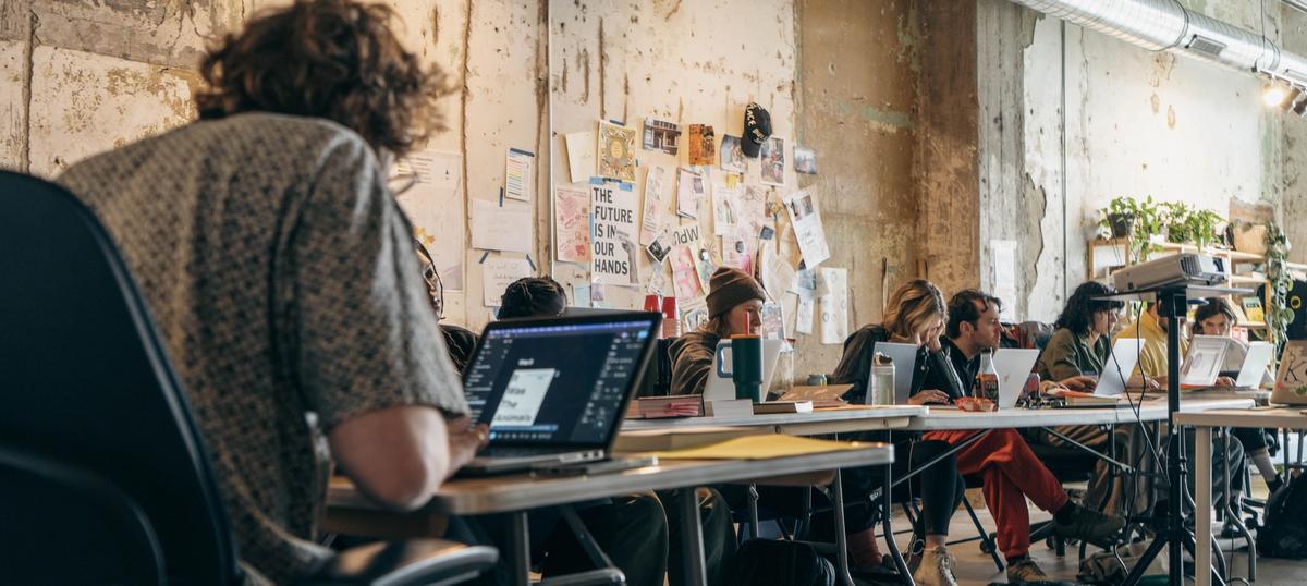 A group of people working on laptops at a long table in a creative workspace.