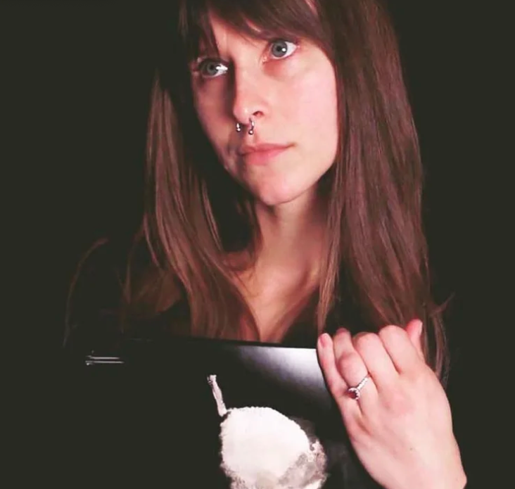 A woman with brown hair and a septum piercing holds a black object with a white graphic.