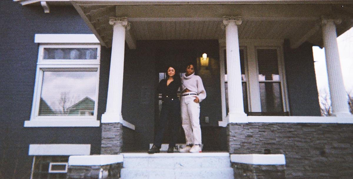 A woman in black and another in cream stand on the porch of a dark grey house.