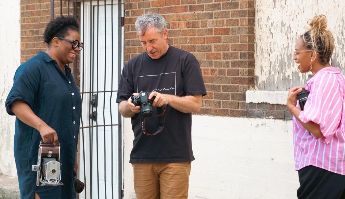 Three smiling people outdoors with cameras; a man checks a digital camera while a woman holds a vintage camera.