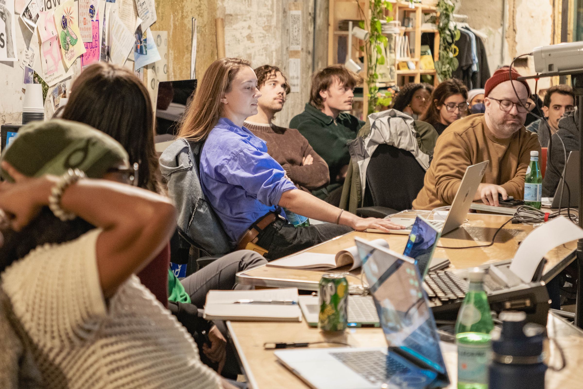 A group of people seated at long tables with open laptops, attending a workshop or presentation.