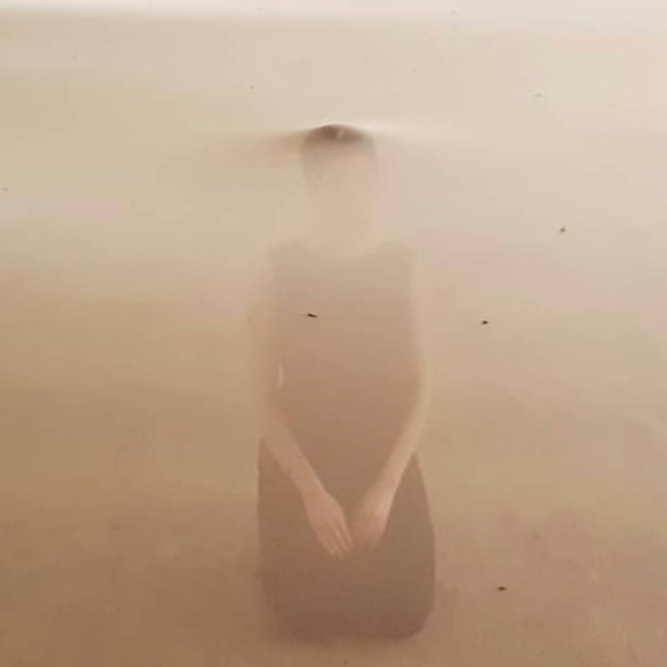 A kneeling figure in a dark dress, almost submerged in a light brown, hazy medium.