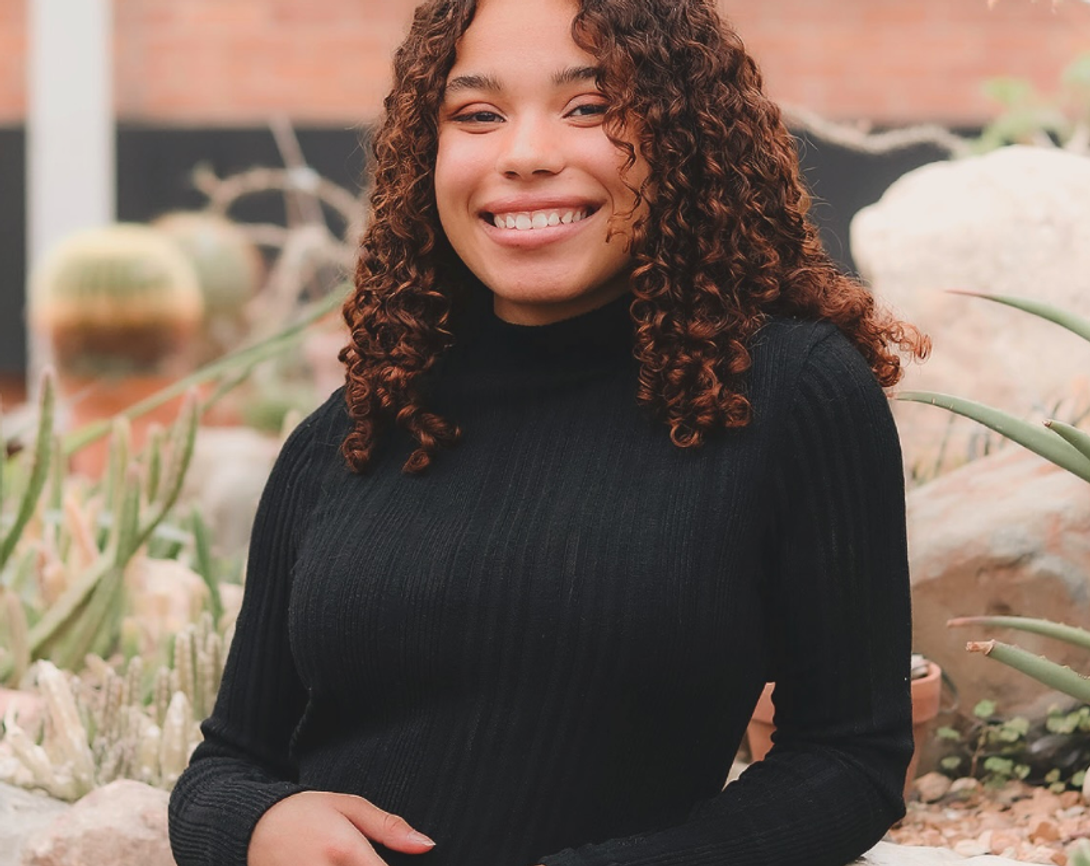 Smiling young woman with curly hair in a black turtleneck, surrounded by cacti and succulents.