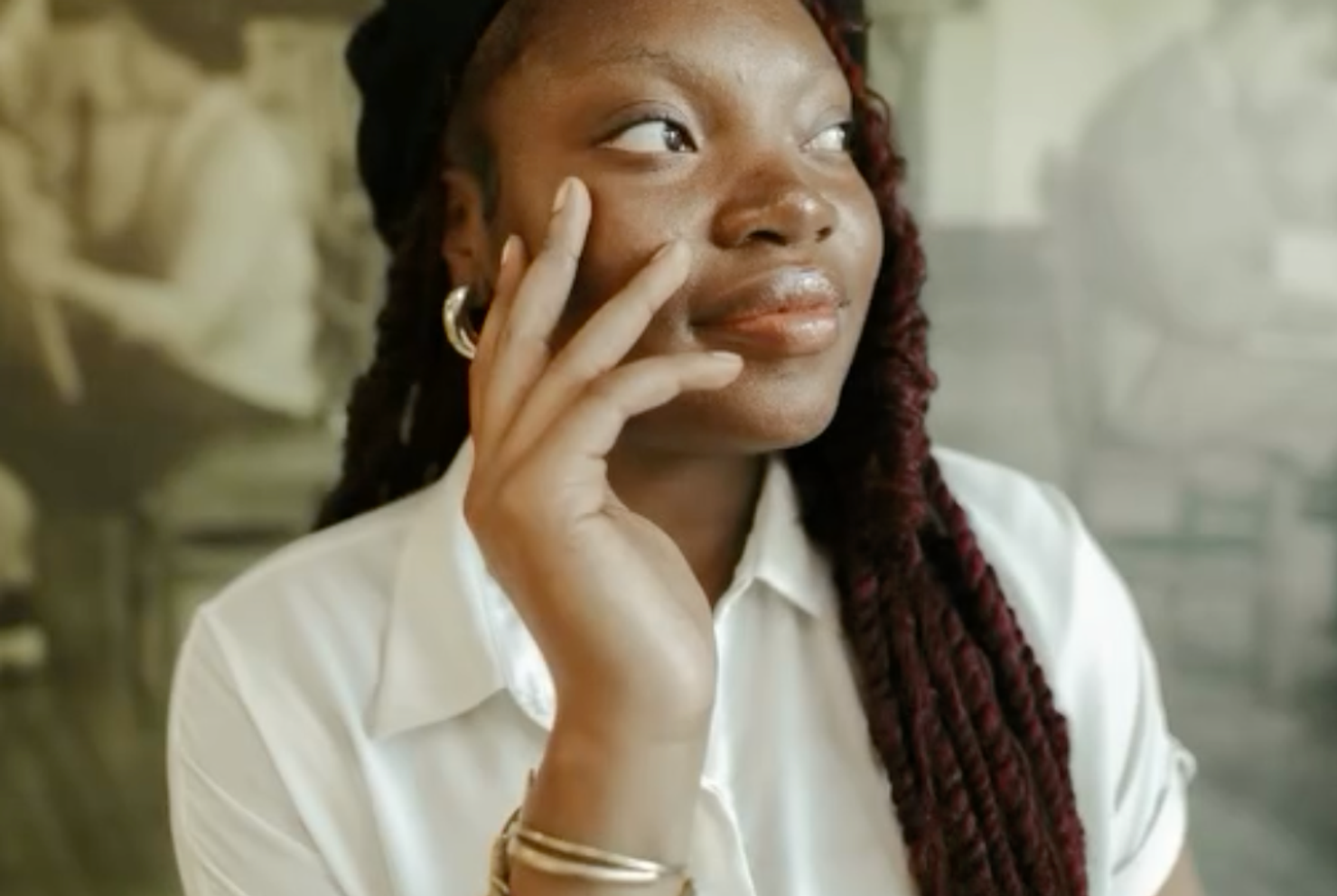 Young Black woman in a black beret and braids looks thoughtfully to the side, hand to her face, against a blurred vintage photo background.