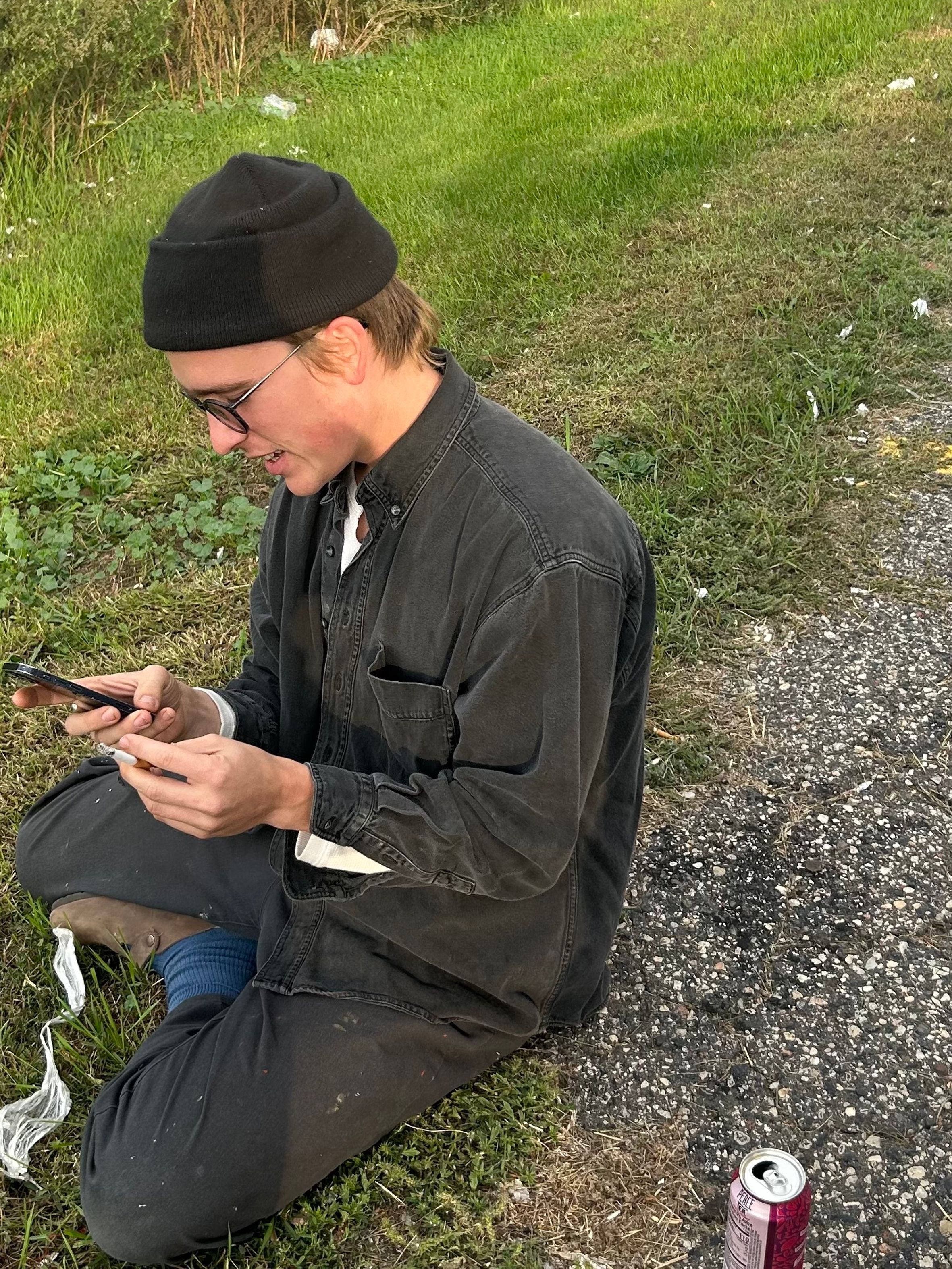 A young man in a black beanie and glasses sits cross-legged on the grass by an asphalt path, looking at his phone with a field in the background.