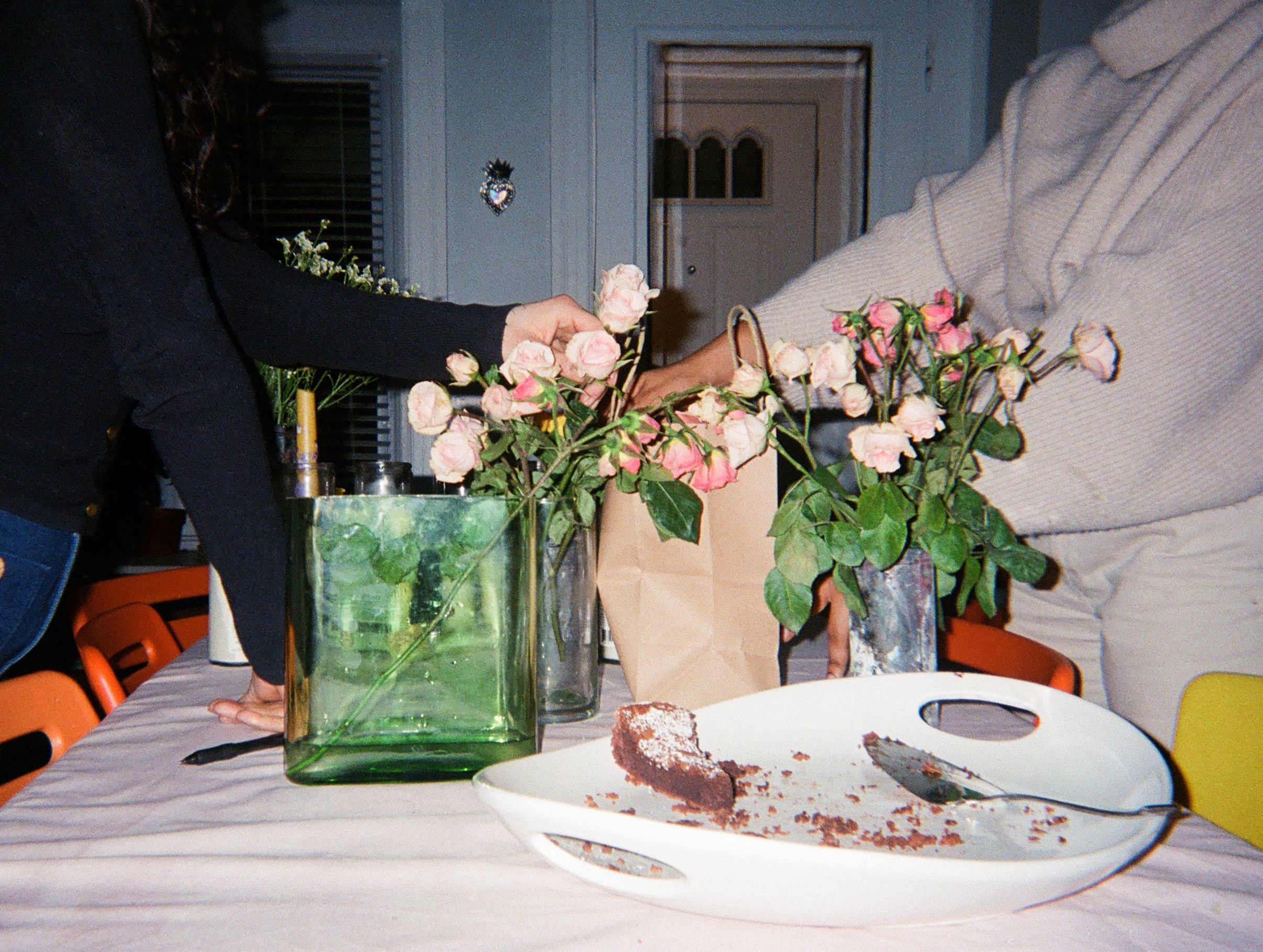 Two people arranging pink roses in vases on a table, with a partially eaten cake on a white plate.