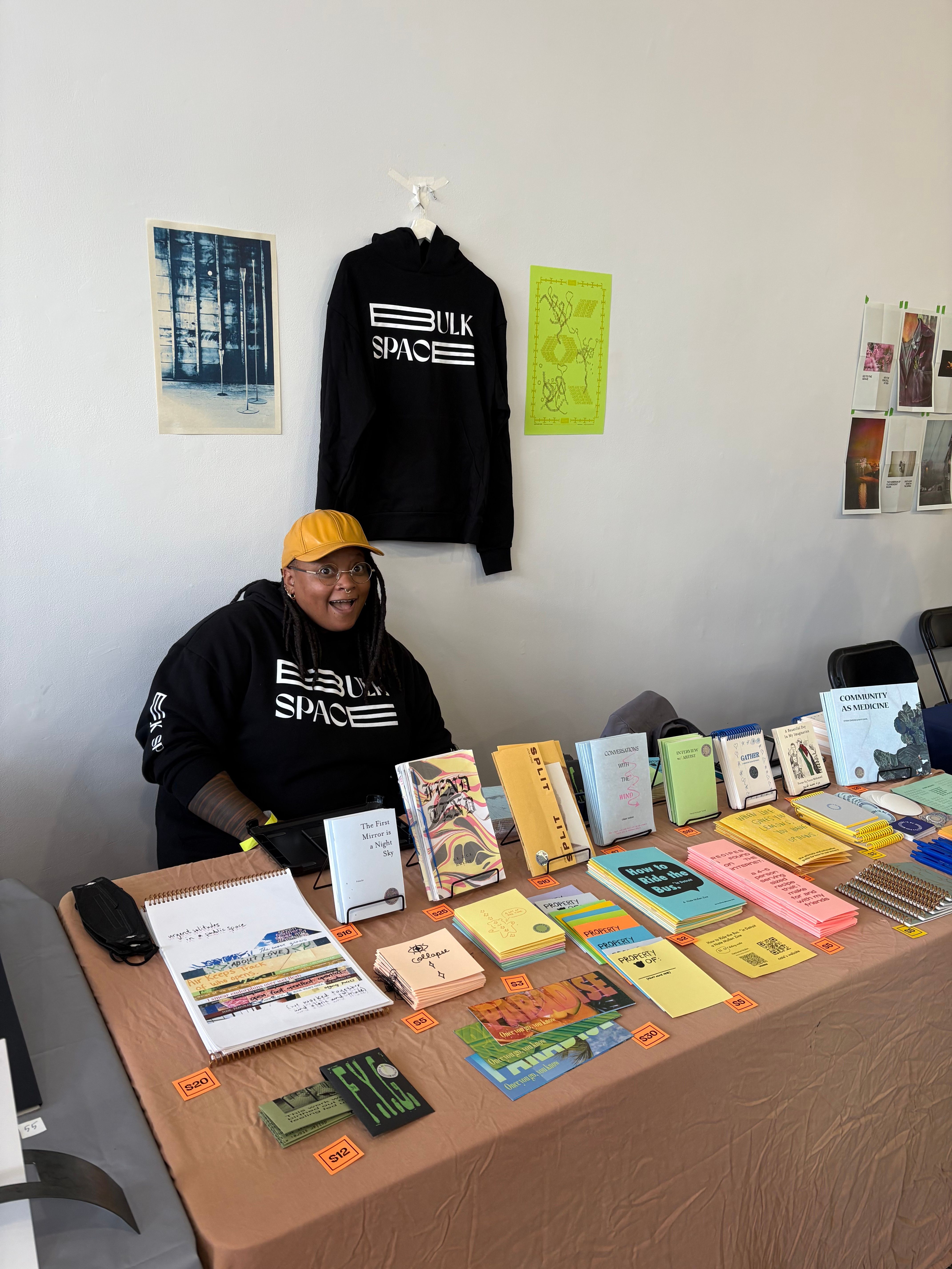 A person in a yellow hat and black "BULK SPACE" hoodie sits smiling behind a table filled with various books and publications.