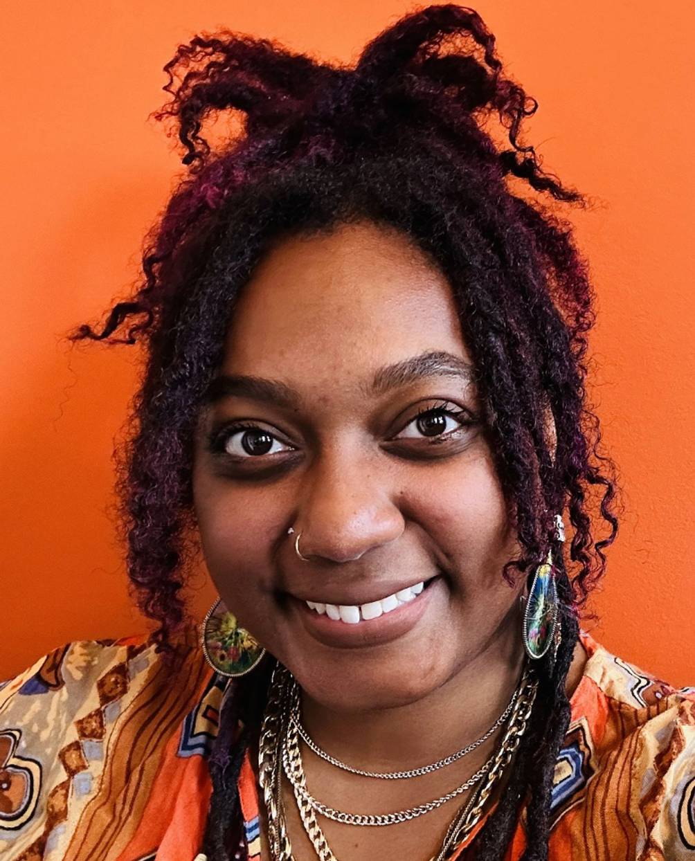 Smiling person with purple dreadlocks, layered necklaces, and a patterned shirt against an orange background.