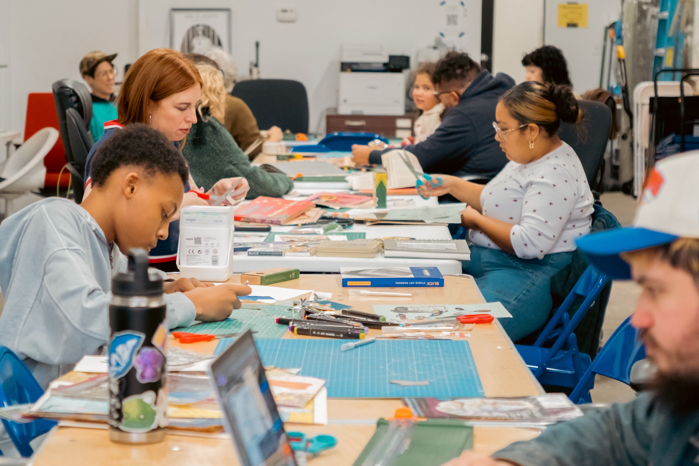 A diverse group of people works on crafts and laptops at long tables in a brightly lit workshop.