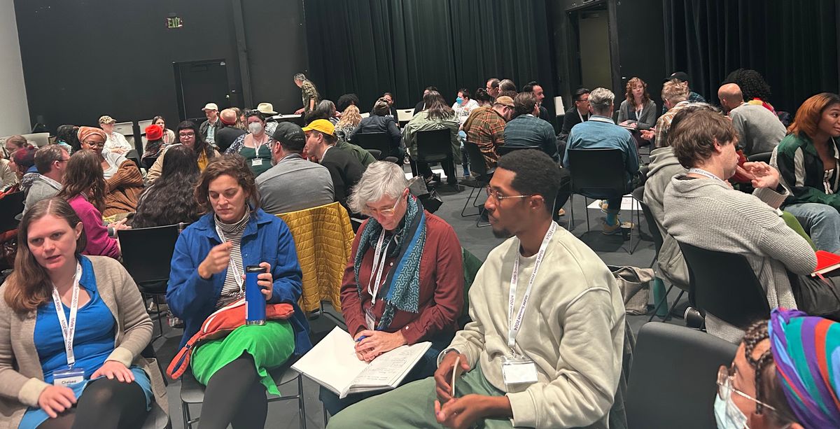 A diverse group of people sit in a large, dark room, attending a workshop or discussion.