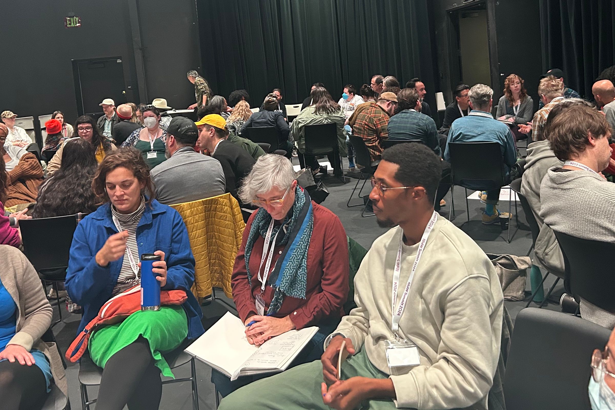 A diverse group of people sit in a large, dark room, attending a workshop or discussion.