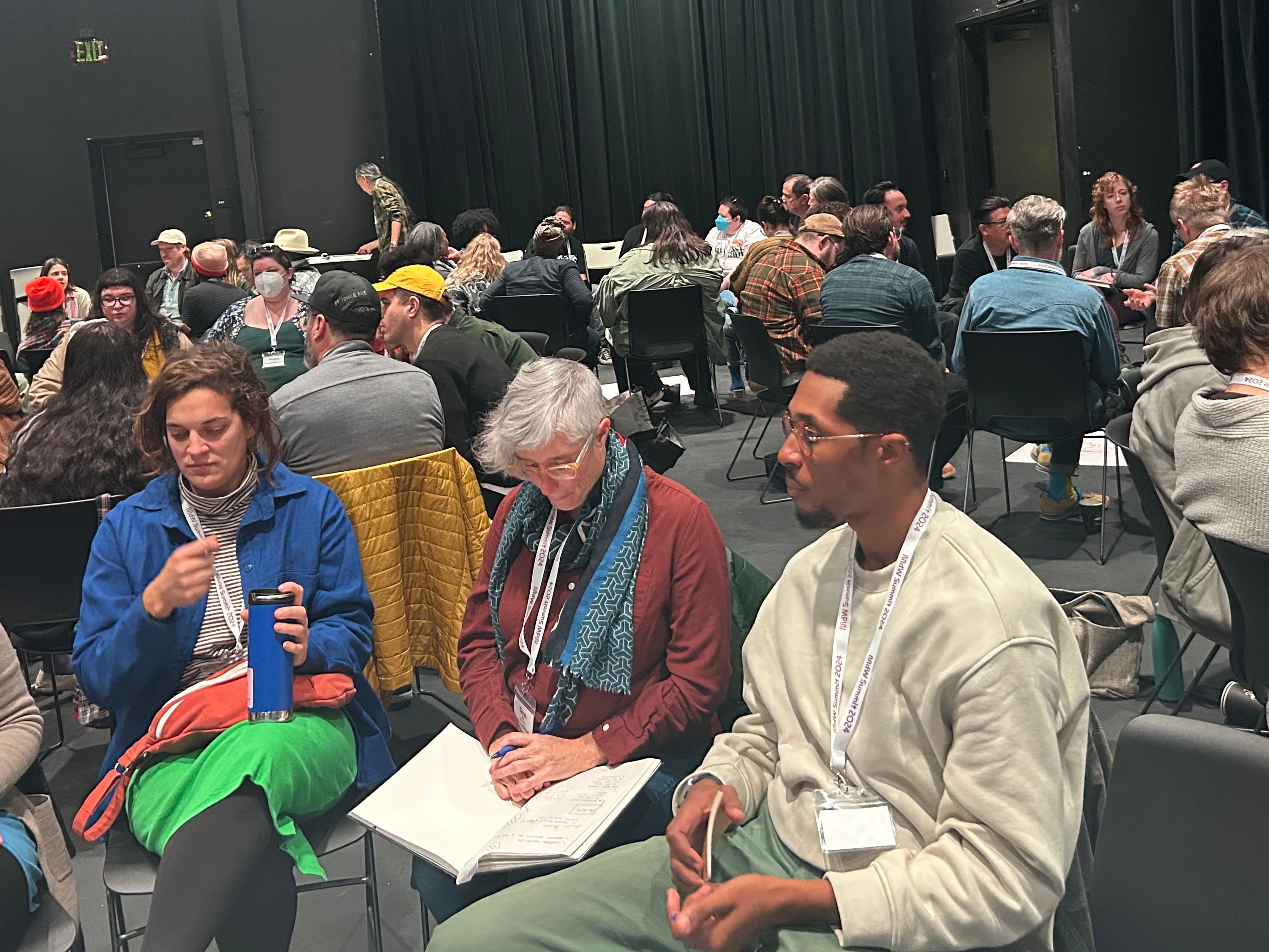 A diverse group of people sit in a large, dark room, attending a workshop or discussion.