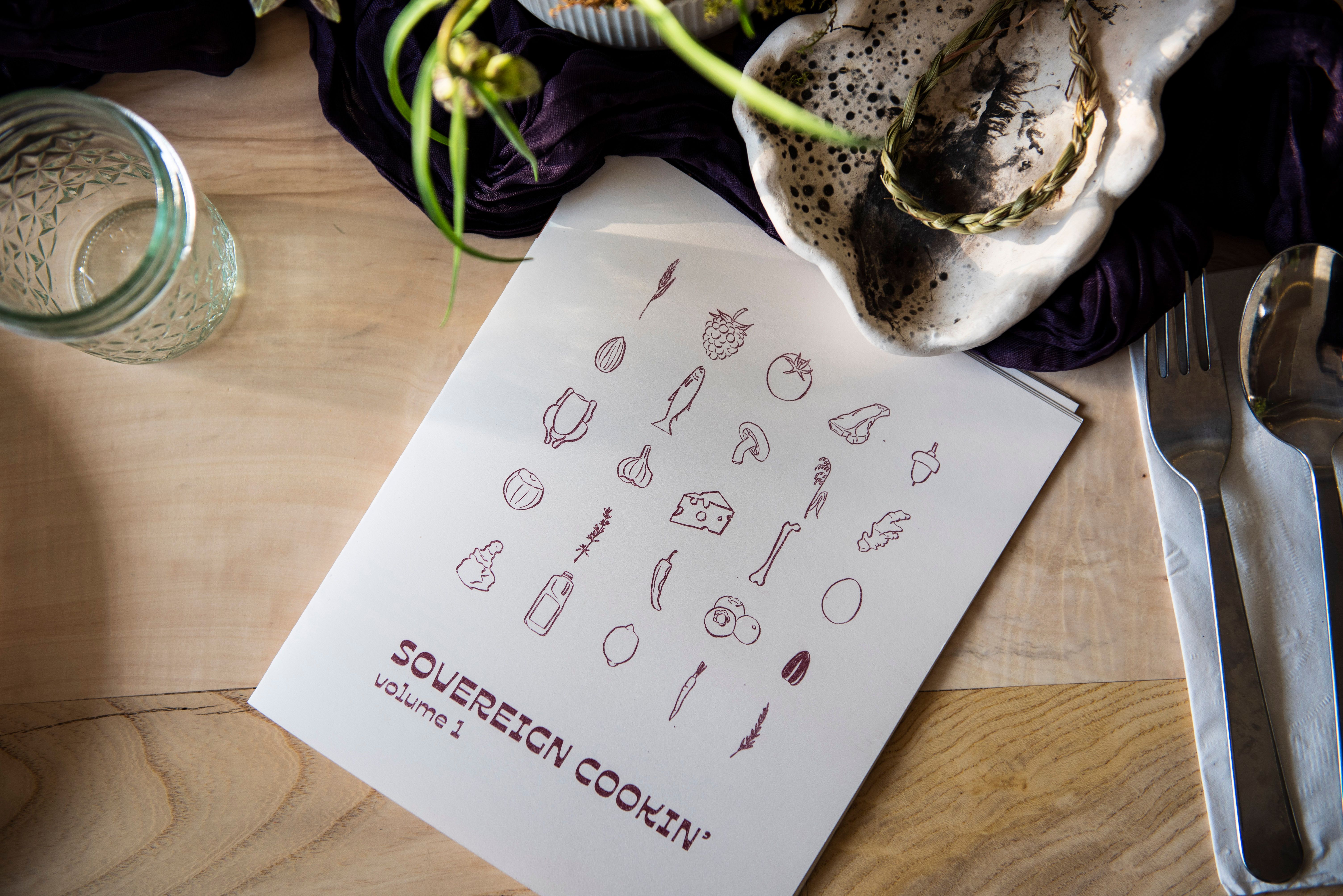A white cookbook titled "SOVEREIGN COOKIN' volume 1" featuring food illustrations, placed on a wooden table with a glass jar and silverware.