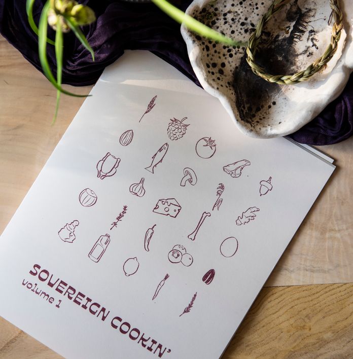 A white cookbook titled "SOVEREIGN COOKIN' volume 1" featuring food illustrations, placed on a wooden table with a glass jar and silverware.