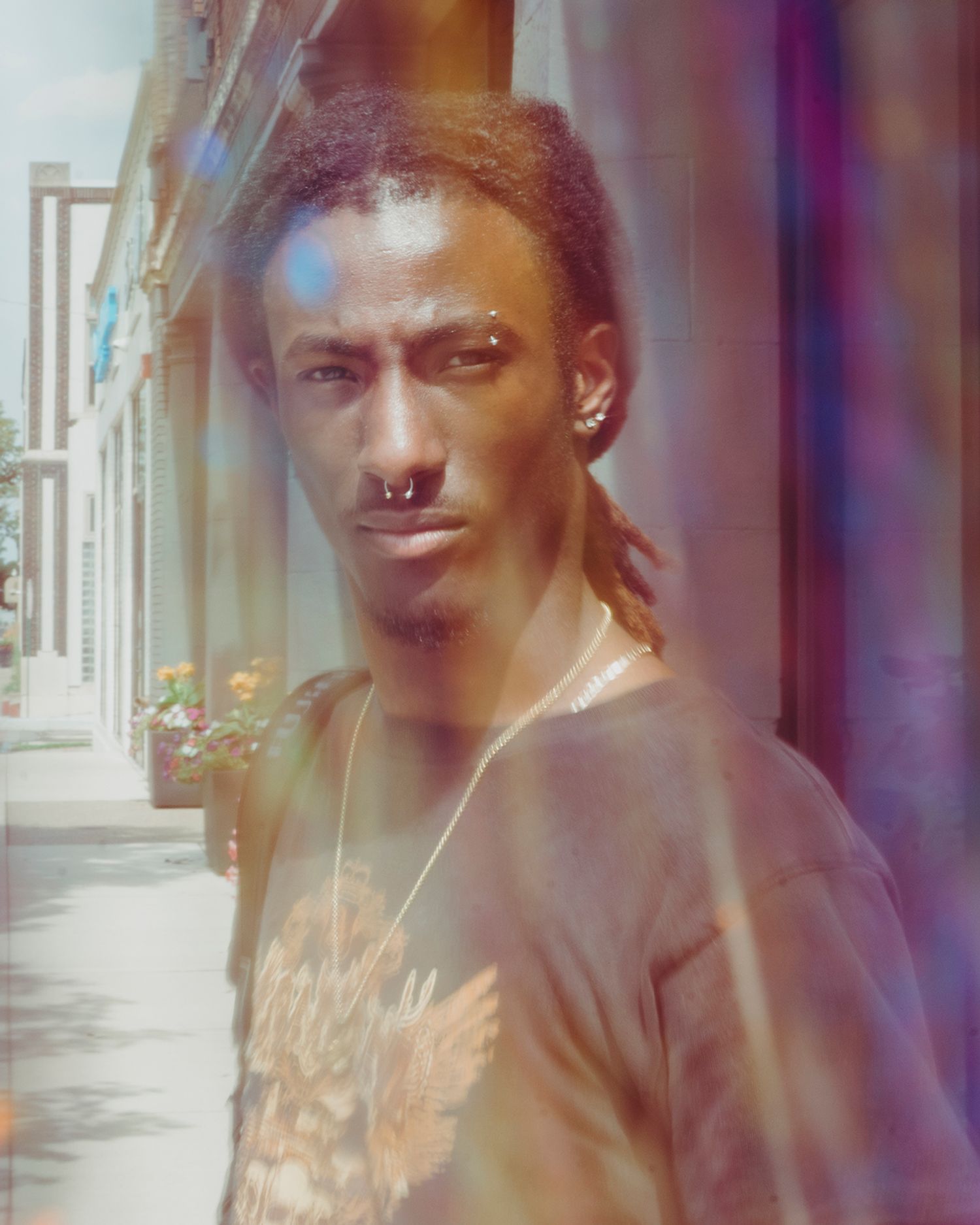 A person with dreadlocks, nose ring, and eyebrow piercing looks forward, seen through colorful lens flares and a soft haze.