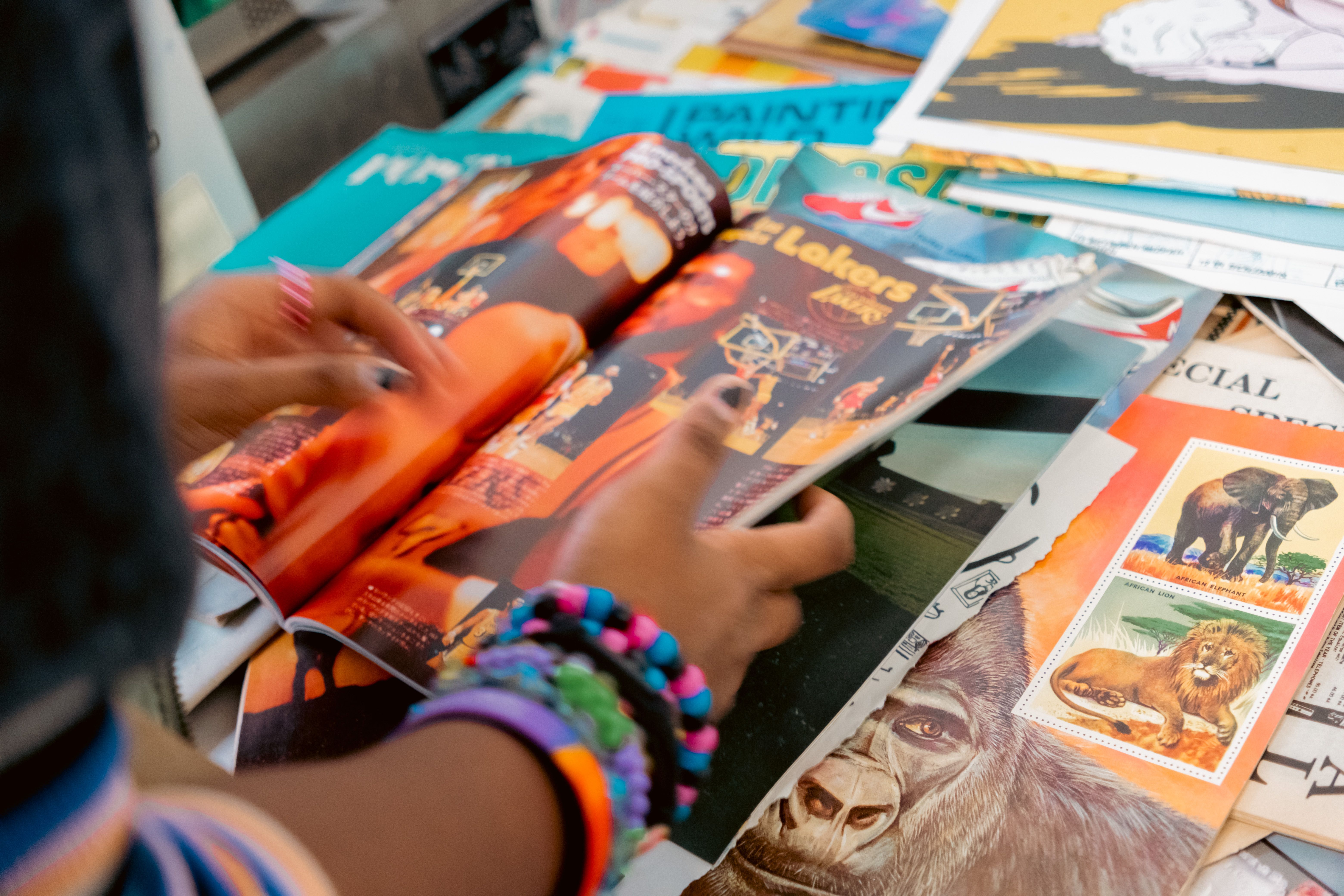 Hands wearing colorful bracelets flip through a magazine, with "Lakers" visible on the page, surrounded by other publications.