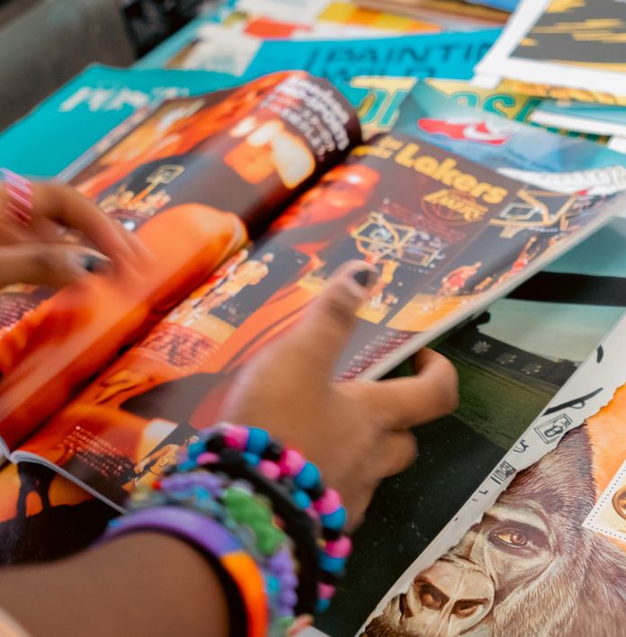 Hands wearing colorful bracelets flip through a magazine, with "Lakers" visible on the page, surrounded by other publications.