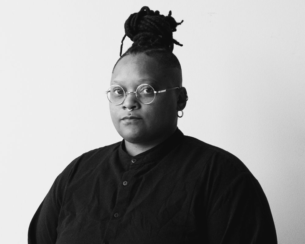 A person with dark skin, glasses, nose and lip rings, and dreadlocks in a bun with shaved sides, wears a dark shirt in a black and white portrait.