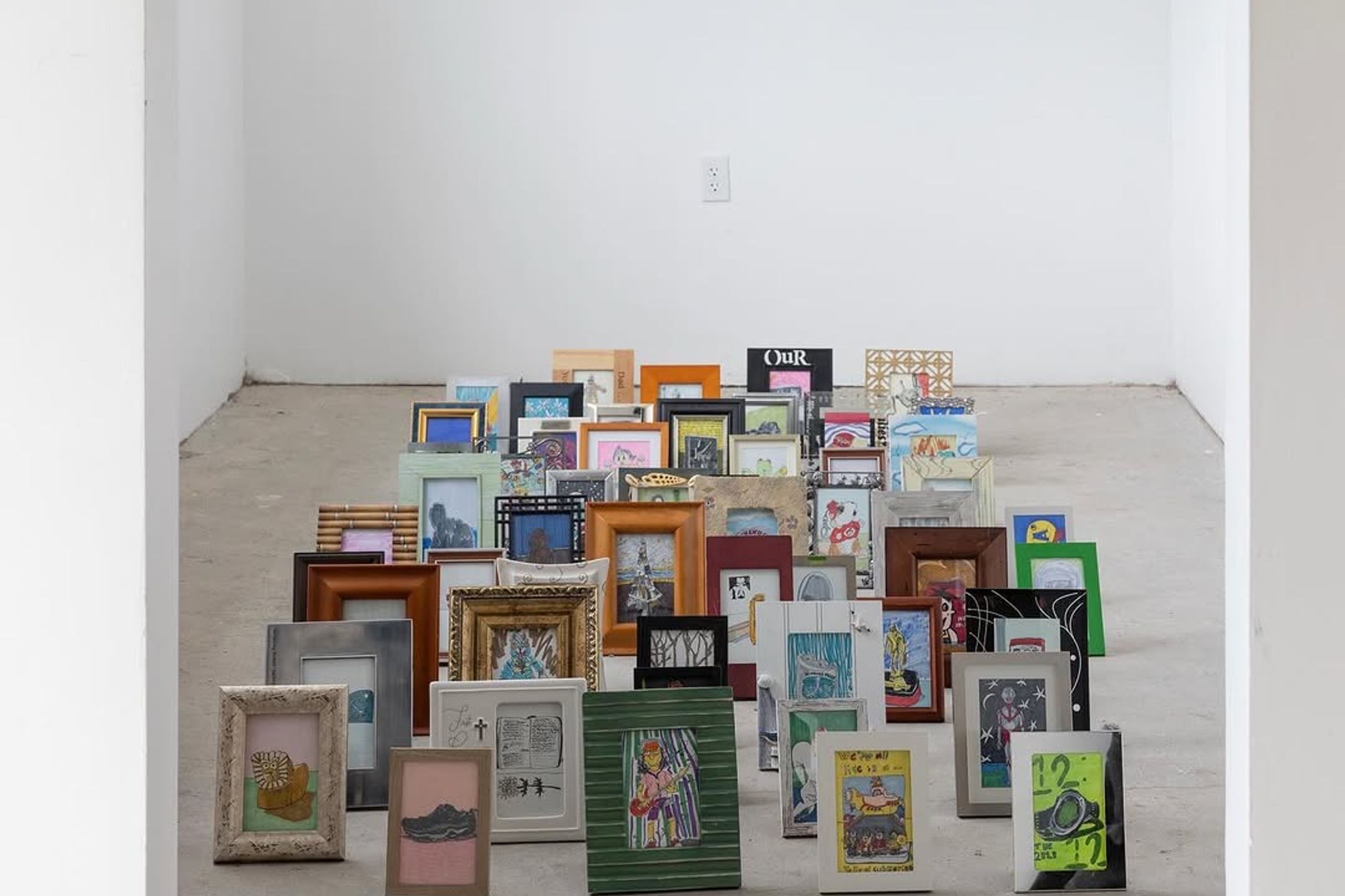 A large collection of diverse framed pictures and artworks are arranged in a rising formation on a concrete floor in a minimalist white room.