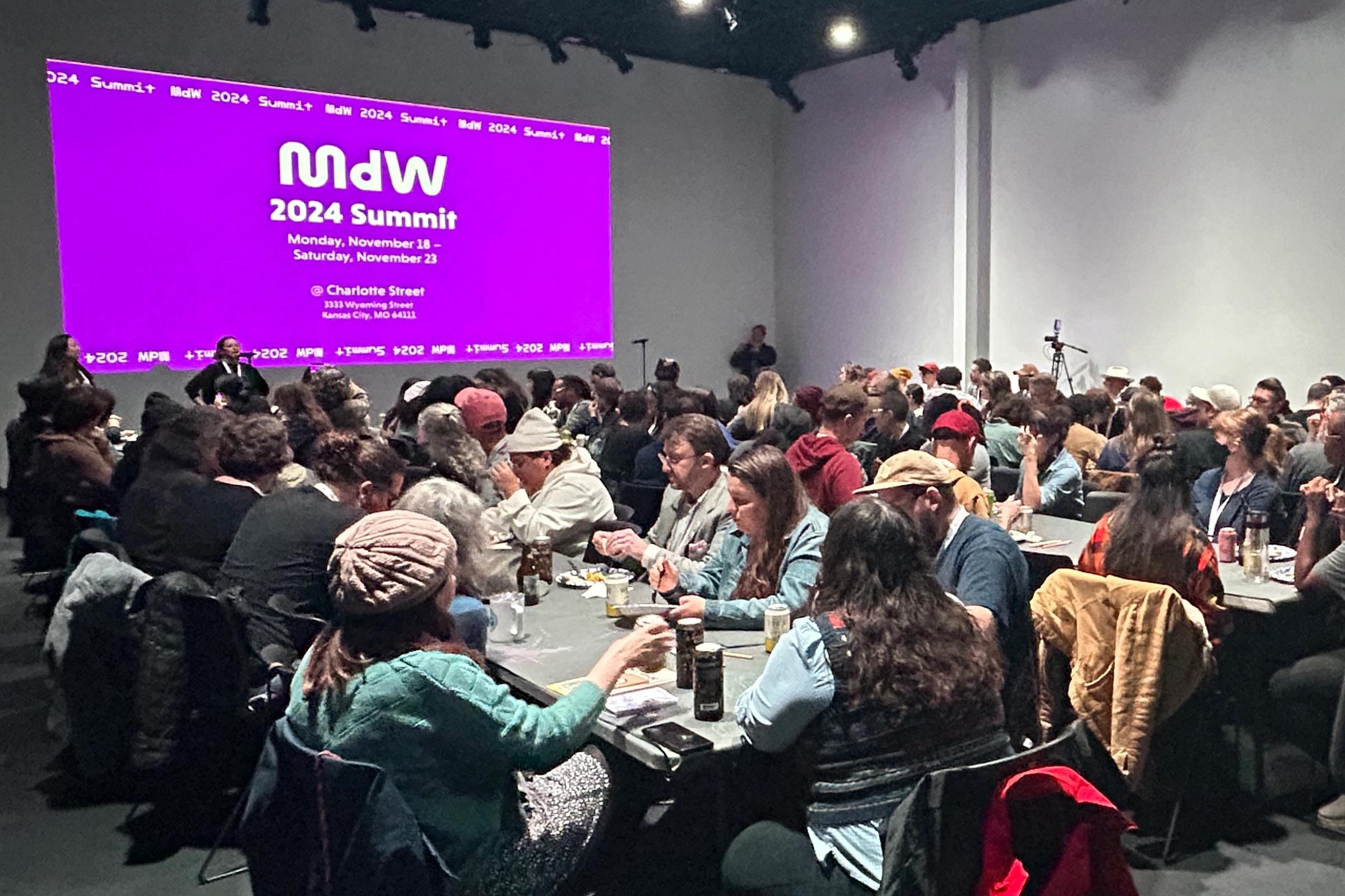 A large group of people seated at tables in a room, facing a screen displaying "MDW 2024 Summit" on a purple background.