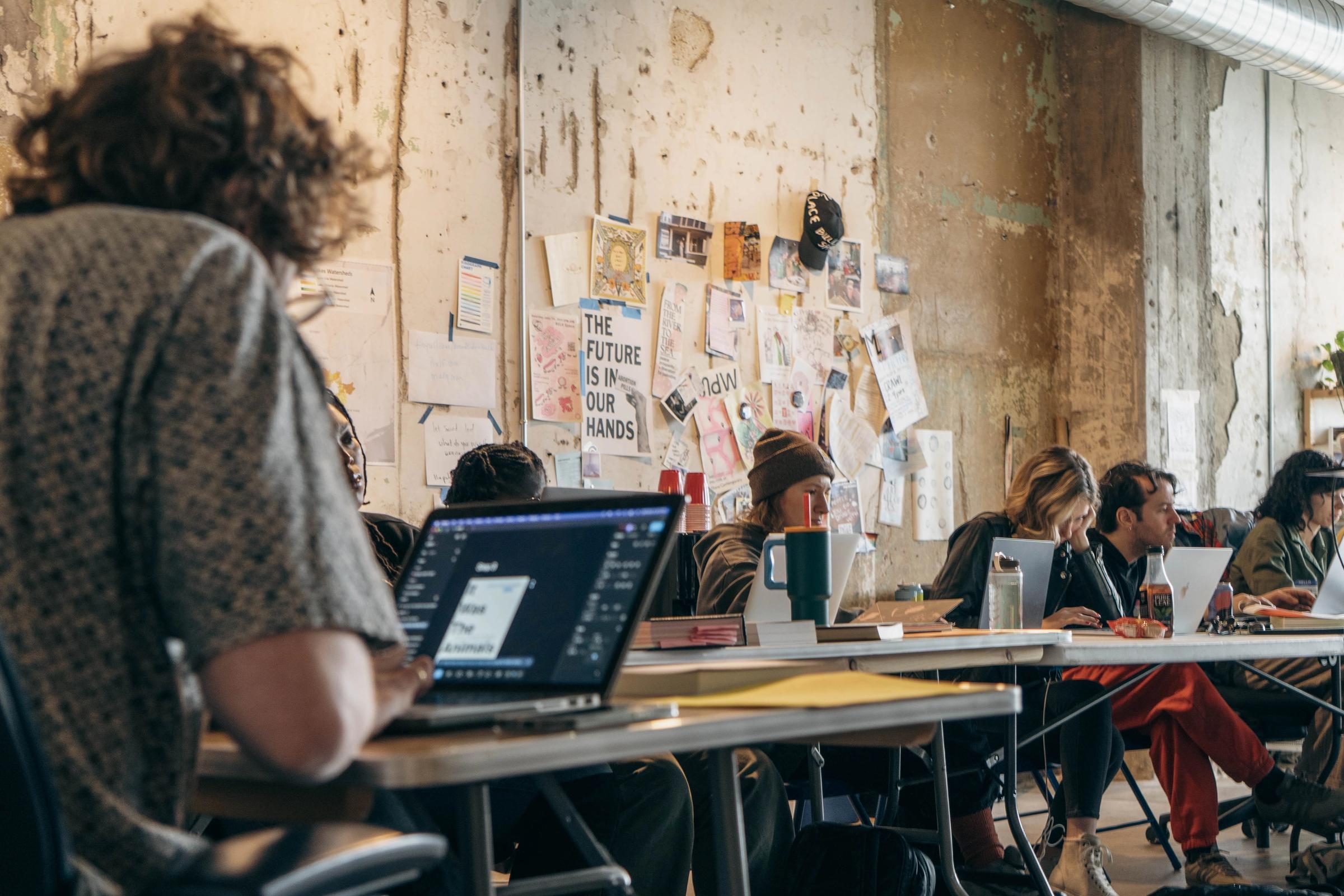 People work on laptops at tables in an industrial-style room with notes on the wall.