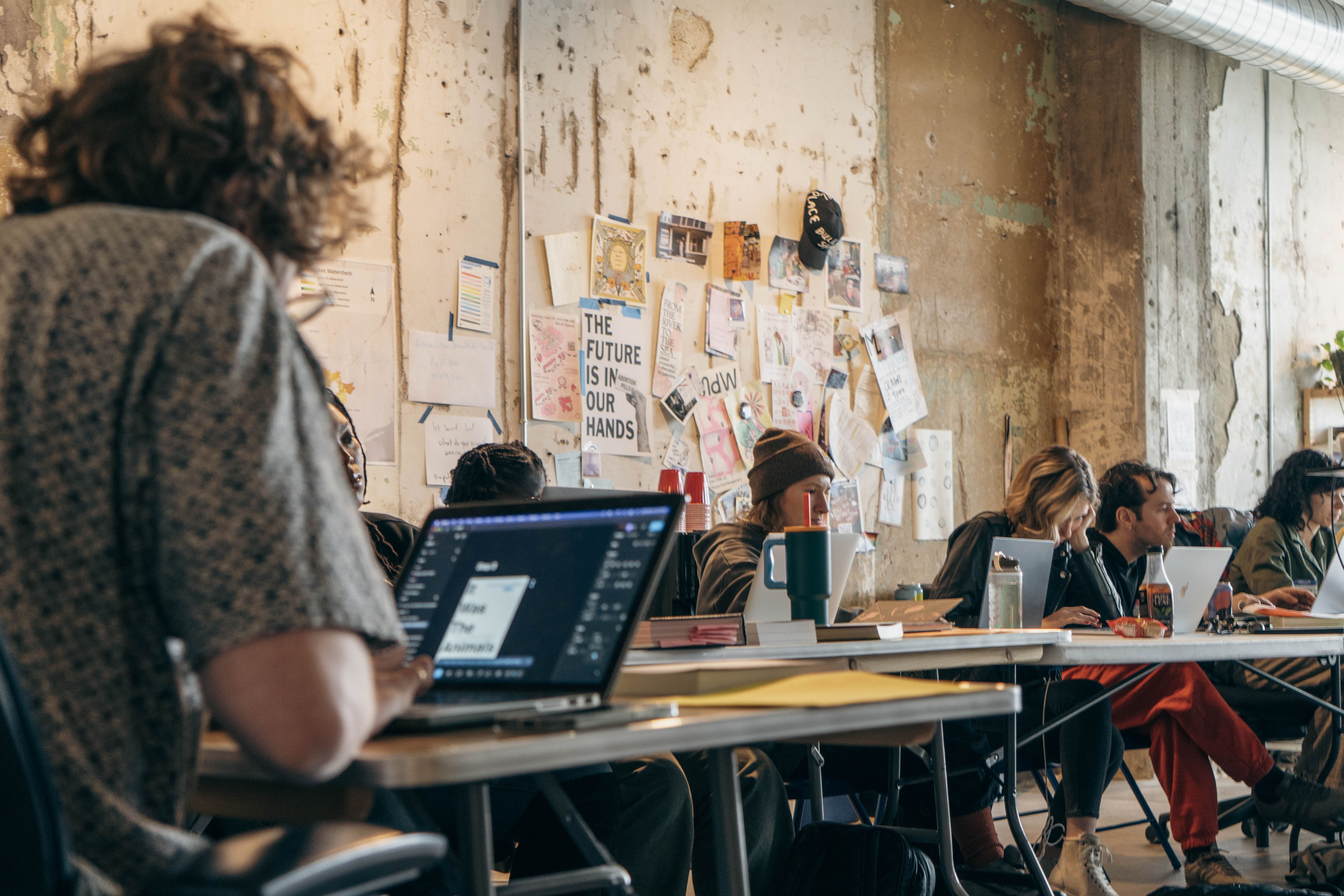 People work on laptops at tables in an industrial-style room with notes on the wall.