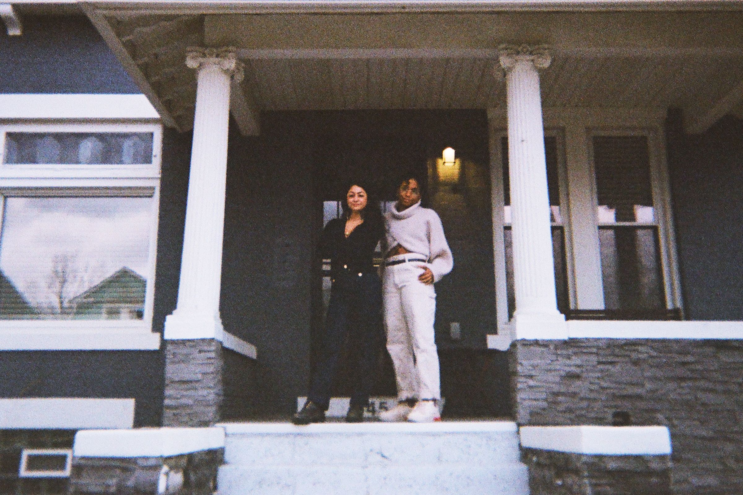 A woman in black and another in cream stand on the porch of a dark grey house.