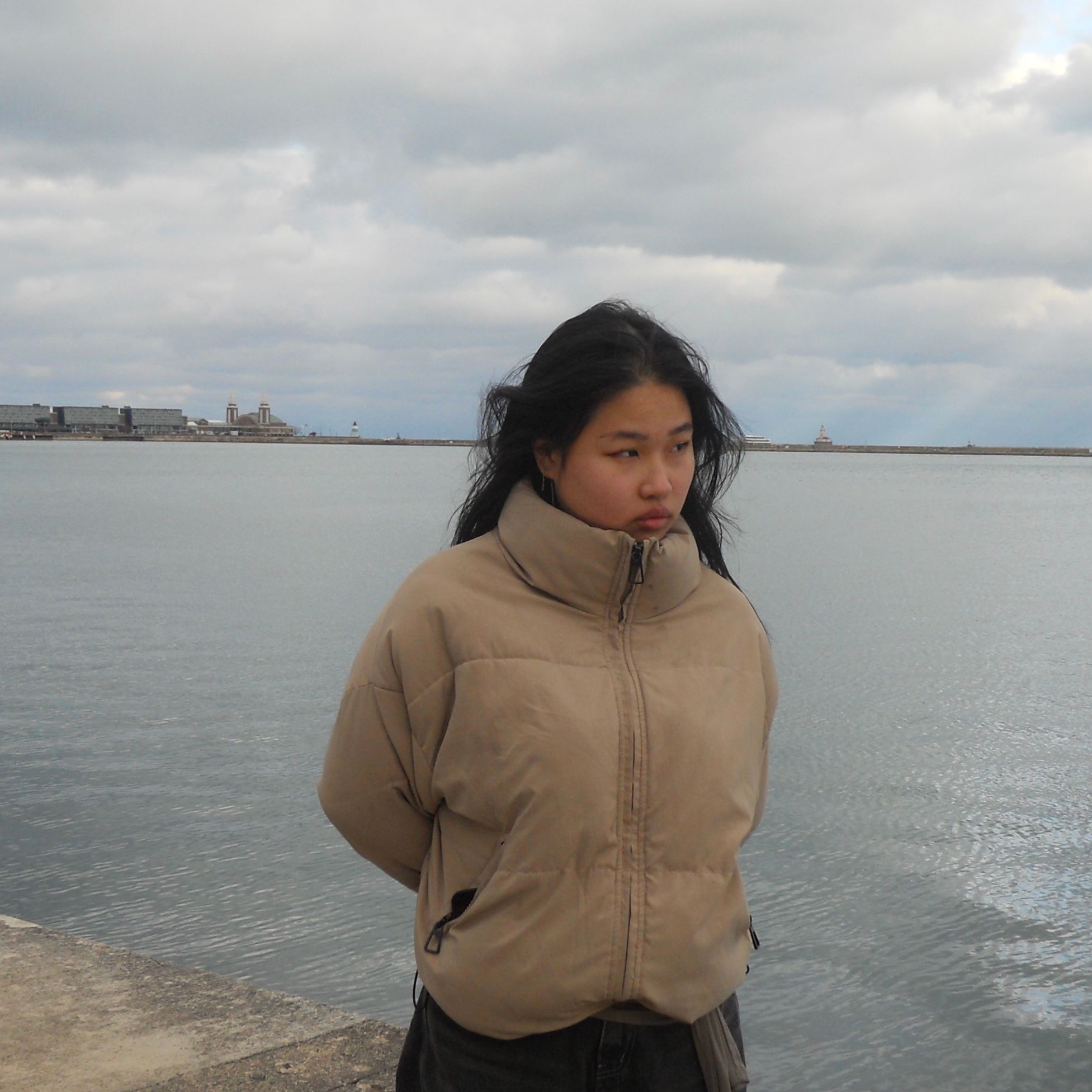 A young person in a tan puffer jacket stands by a cloudy lake with distant buildings and a lighthouse.