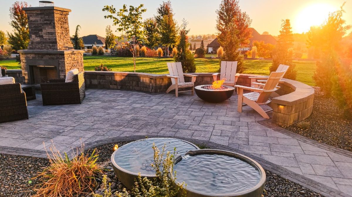 Golden-hour view of the Caldwell patio with a stone fireplace, fire pit, pavers, and Aquascape fountain