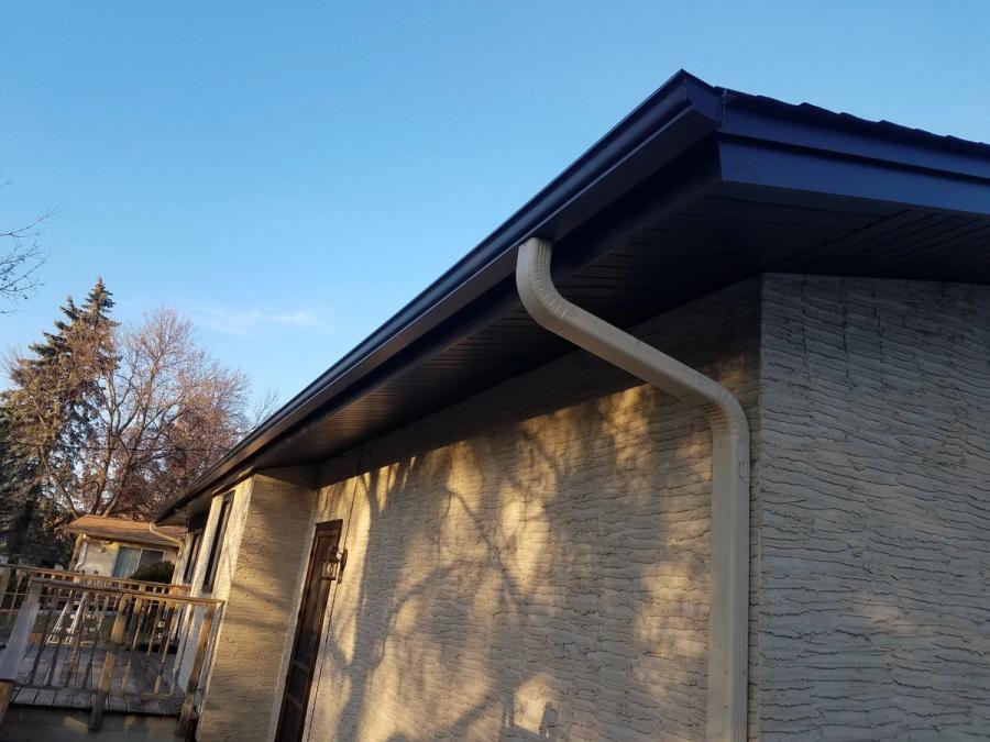 Dark gutter along a home’s roofline with a light-colored downspout beside the exterior wall.