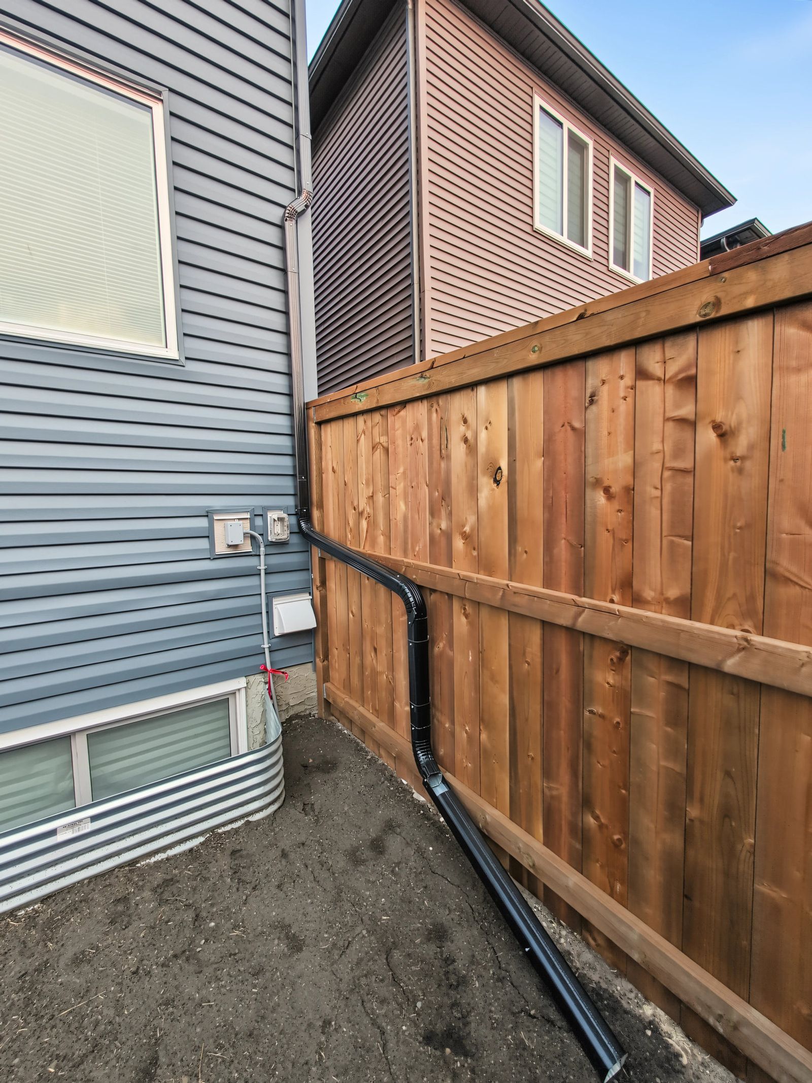 The downspout extension running along the base of the fence, carrying roof water away from the garage wall