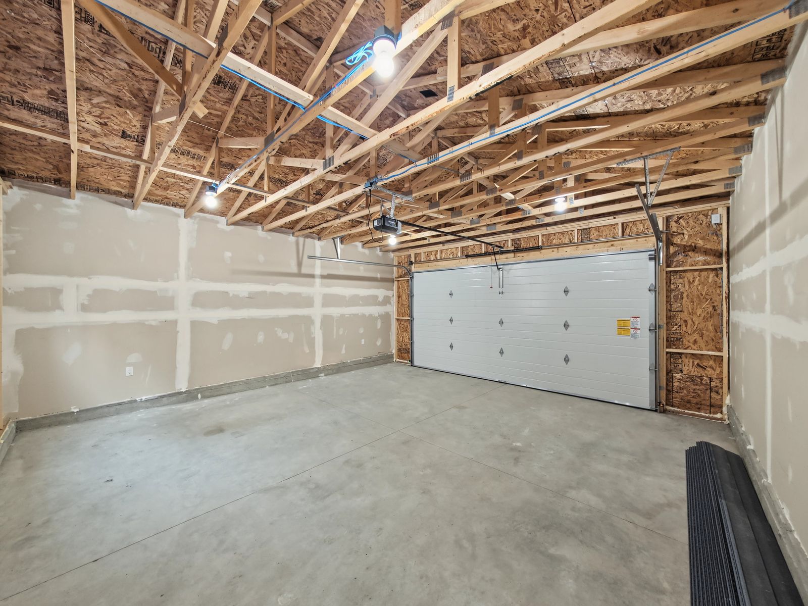 Inside the garage looking at the closed overhead door: one side wall boarded and taped, open trusses above and the chain-drive opener on its ceiling rail