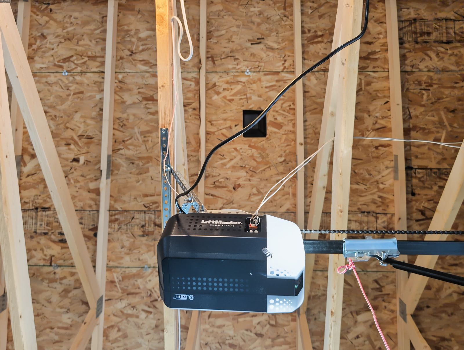 A LiftMaster ceiling-mount opener hanging from the trusses on its chain rail, with the wiring run along the bare OSB sheathing