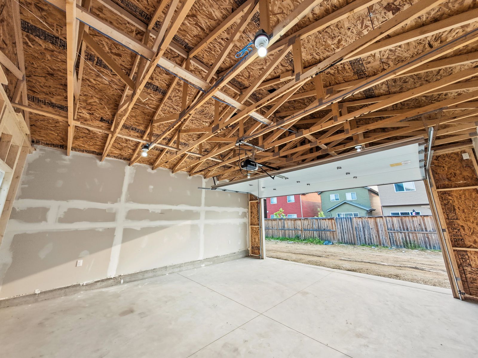 Inside the garage with the overhead door open to the lane, the taped drywall wall on the left and the trusses overhead