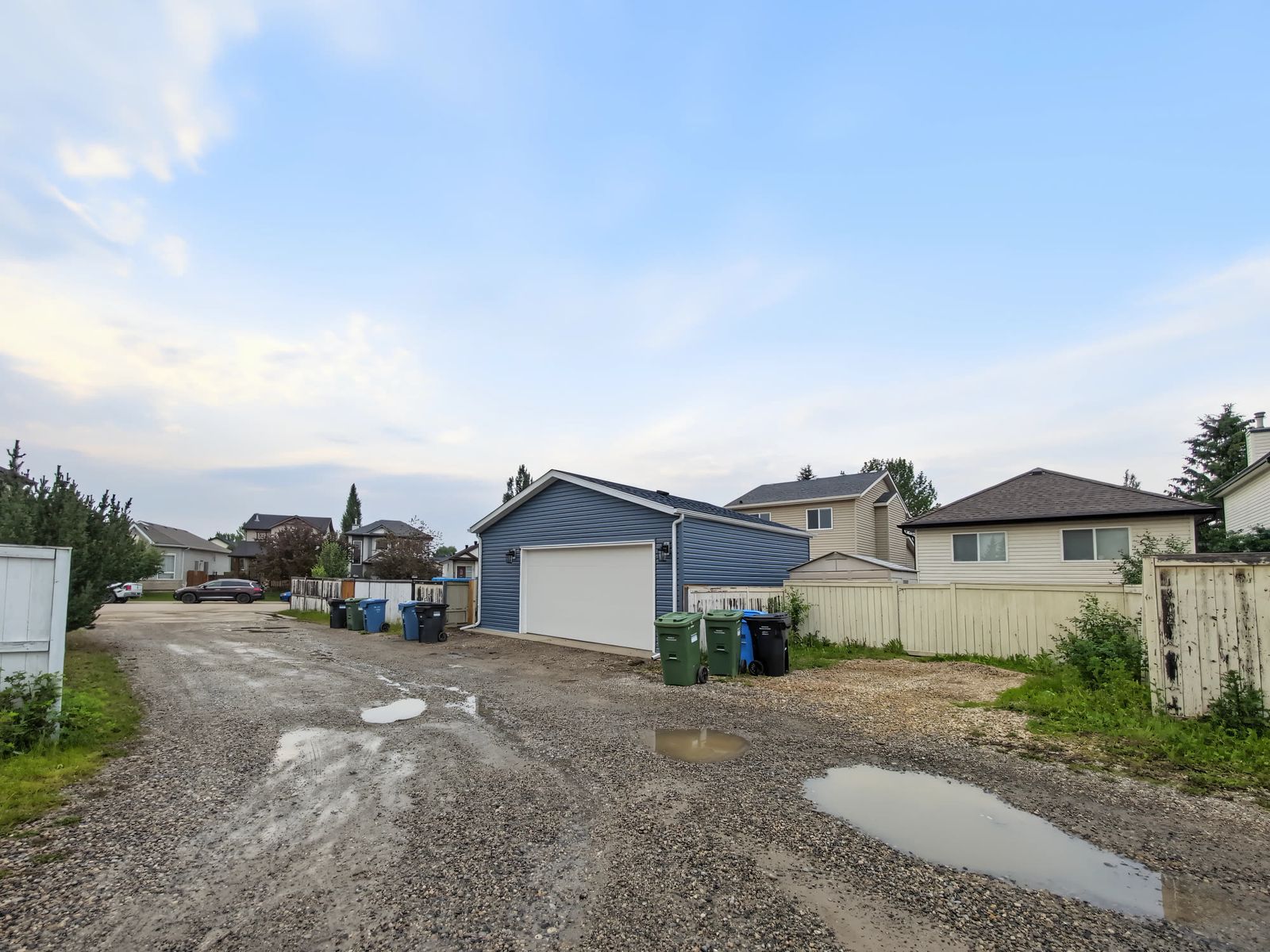 The garage seen along the gravel back lane, blue siding and white overhead door, with bins and fences either side