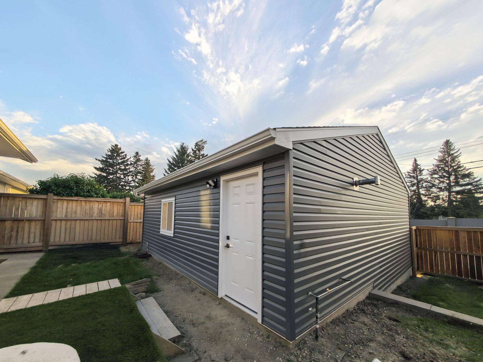 The garage from the yard: white man door and sliding window on the long wall, and on the gable end the heater vent and the gas line coming up out of the ground