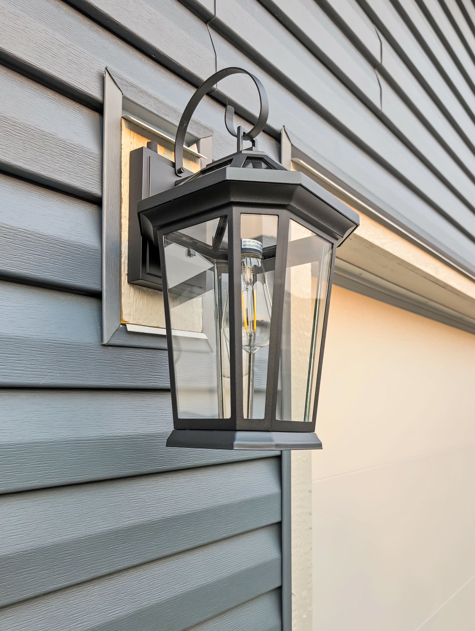 Close view of the black coach lantern on the blue lap siding, lit at dusk, mounted on a white trim block