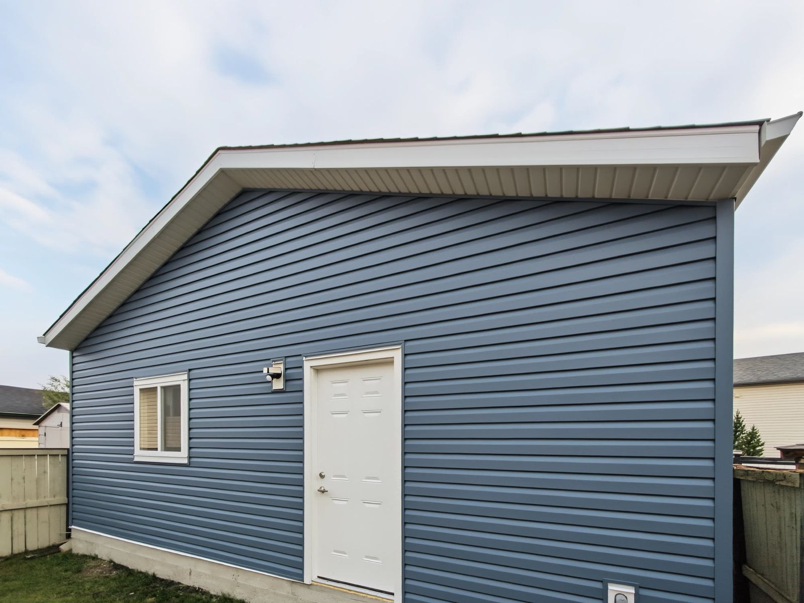 The side of the garage from the yard with the white man door, a wall light above it and the concrete foundation showing below the siding