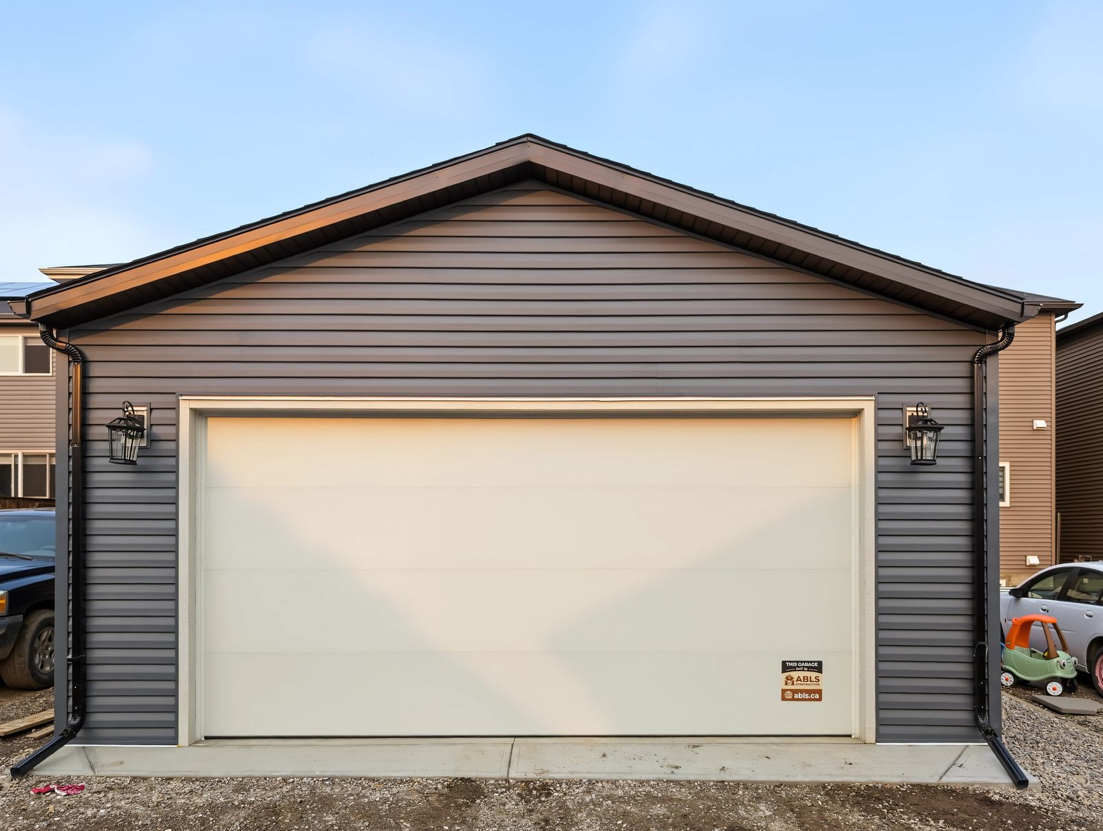 The finished garage straight on from the lane: dark grey-blue siding, a cream flush-panel overhead door in white trim and a lantern each side