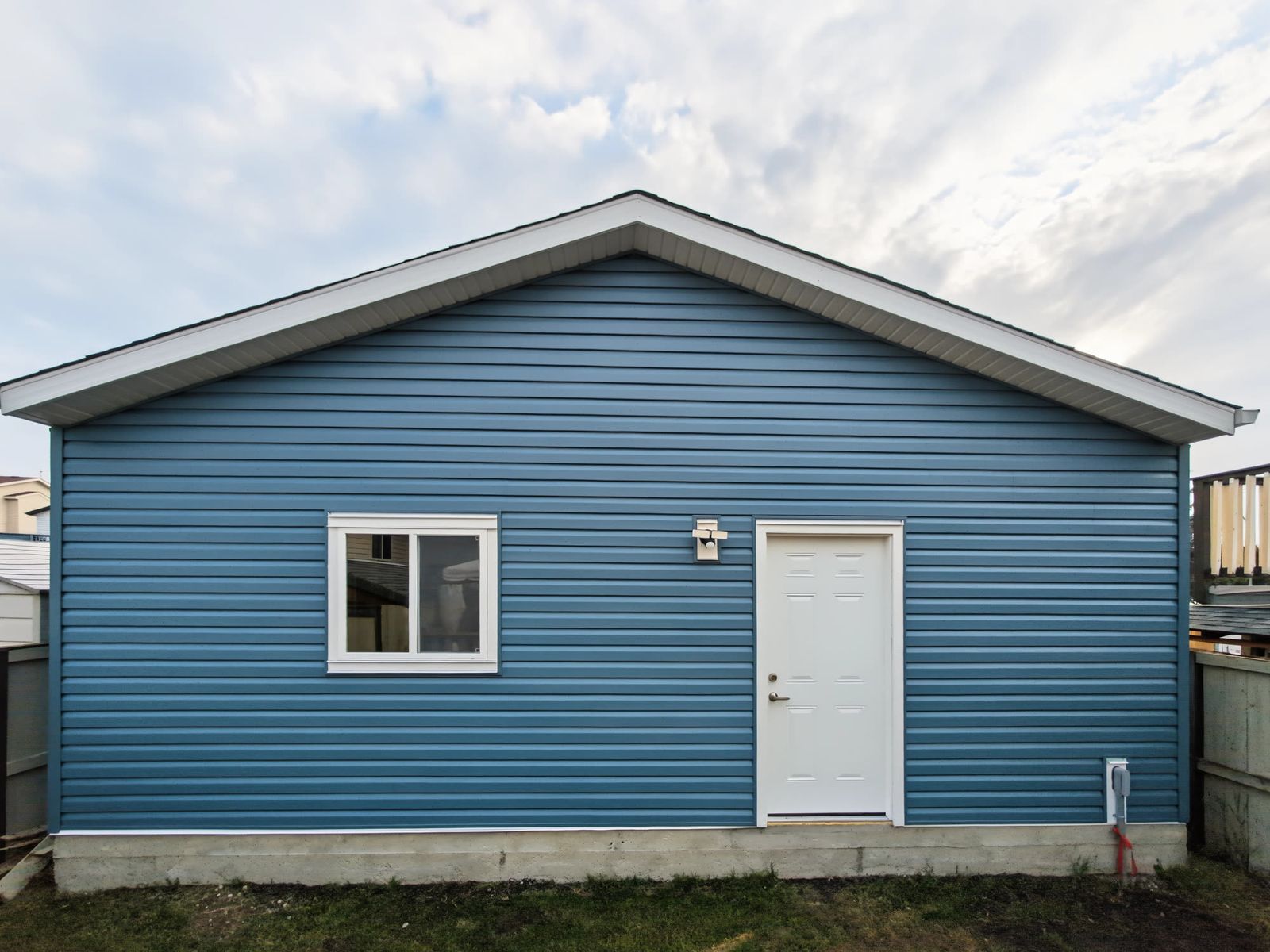 The gable end of the garage straight on: blue lap siding, white man door, a sliding window and an exterior receptacle low on the wall
